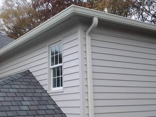 Vertical downspout and horizontal gutter installed on a house with light siding and a shingled roof
